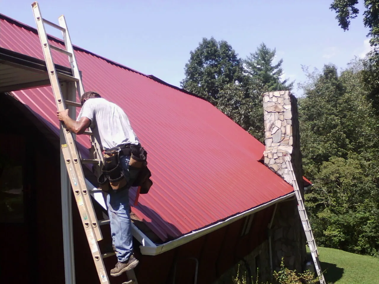Roofer on a ladder beside a red metal roof and stone chimney — Soffit & Fascia Repair by Roofing Brothers in Browntown